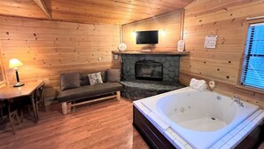 A cozy cabin interior with a stone fireplace, a large bathtub, a sofa, and a television. - BM #9 · Peaceful, Unique Hot Tub in Kitchen - Bear Manor#9 (Big Bear Lake)