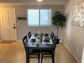 A dining room with a black table set for four, surrounded by chairs, a window with blinds, a door, and a plant.