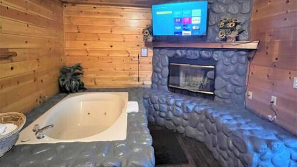 Une salle de bain rustique, aménagée dans une cabane en rondins, avec un mur en pierre, une grande baignoire, un téléviseur fixé au mur et un plafond en bois.