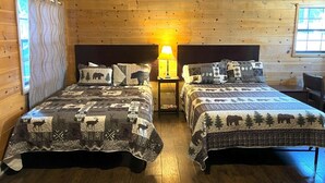 Une chambre de style cabane avec deux lits, chacun recouvert d’une couverture matelassée ornée de motifs animaliers. Une table de chevet avec une lampe se trouve entre les deux lits.