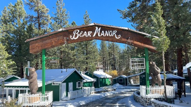 Un arco de madera con la inscripción "Bear Manor", que conduce a una fila de cabañas verdes con barandillas blancas.