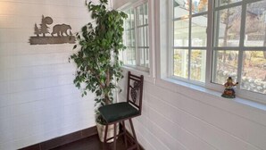 Una habitación con paredes blancas con paneles, techo de madera, un ventanal con vistas a árboles, una silla con cojín verde y un adorno de pared con un oso y árboles.
