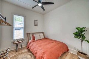 A bedroom with a bed, a ceiling fan, a window, a small table, a chair, and a potted plant.