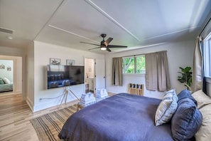Un dormitorio con cama, ventilador de techo, televisor, ventana con cortinas y vista a la vegetación exterior.