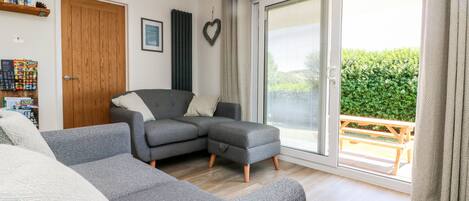 A modern living room with a grey sectional sofa, a wooden door, a framed picture, and a view of a garden through a sliding glass door.