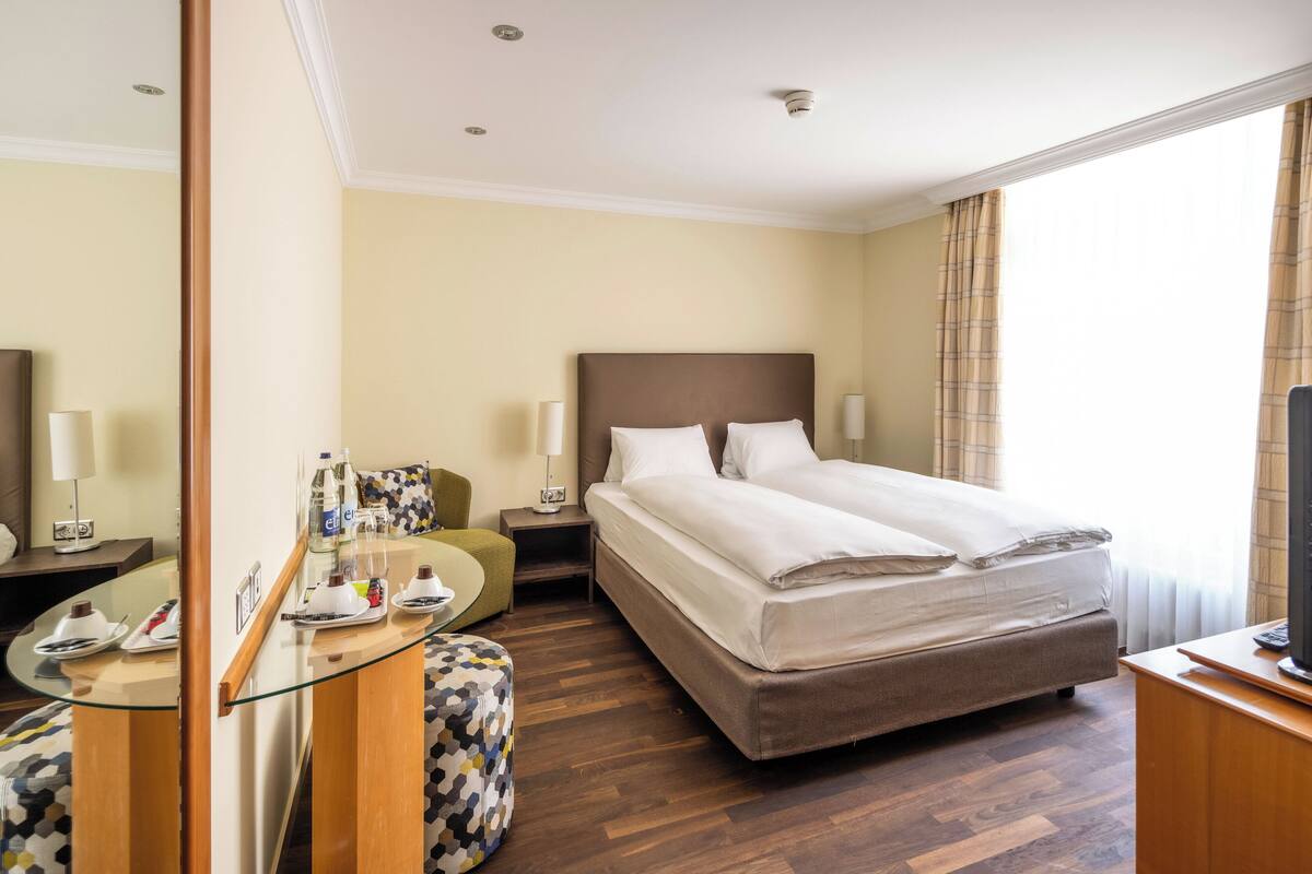 A hotel room with a large bed, a glass table, a chair, a desk, and a television.