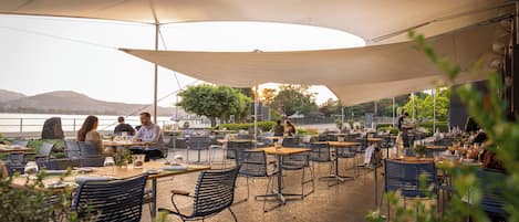 An outdoor dining area with tables and chairs, a large canopy, and a view of the water and hills in the background.