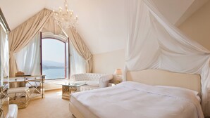 Une chambre avec un grand lit, un coin salon avec une table et des chaises, une vue sur la mer et un lustre.