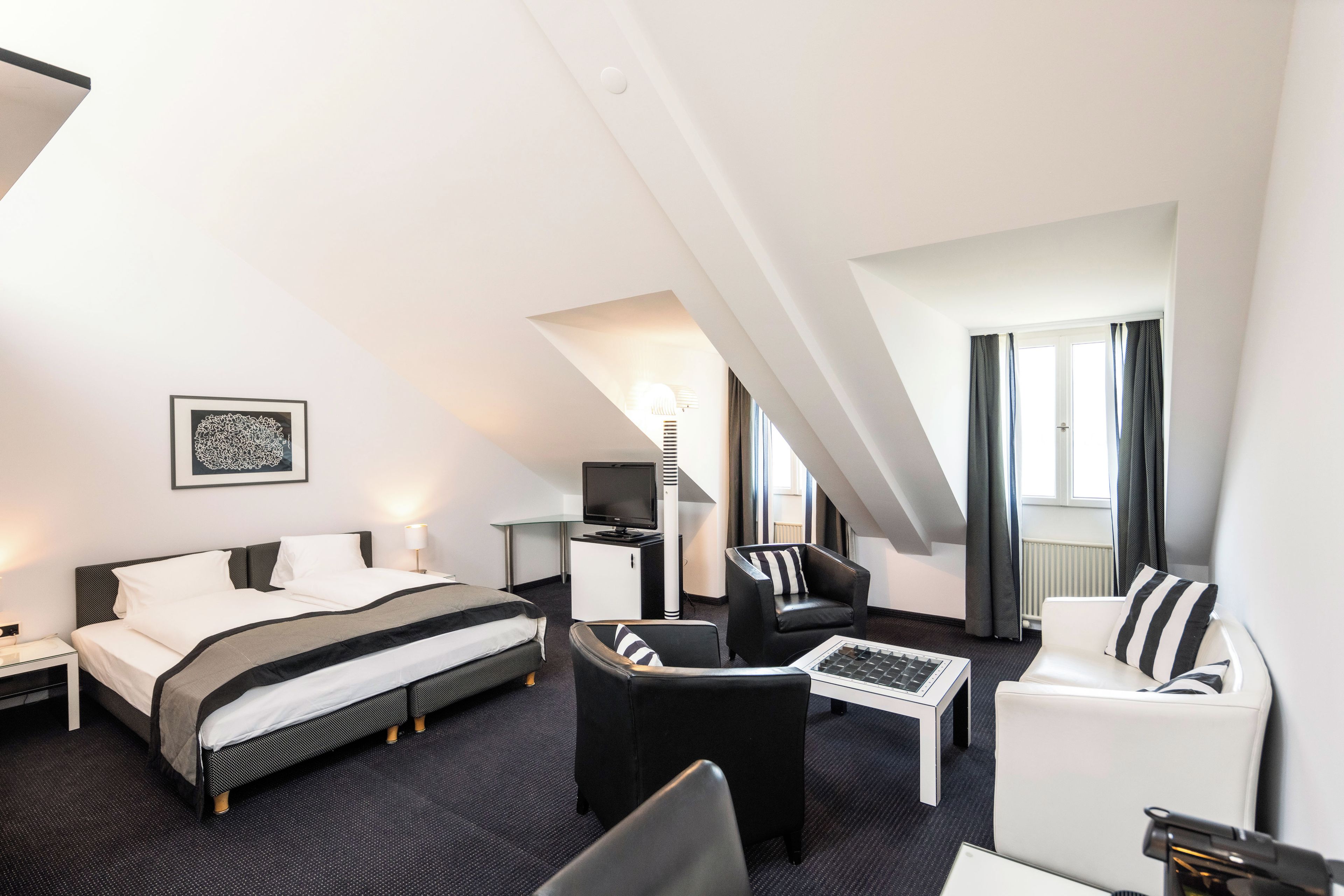 Une chambre d’hôtel moderne équipée d’un lit, de deux fauteuils, d’une table basse, d’un bureau et d’une télévision.