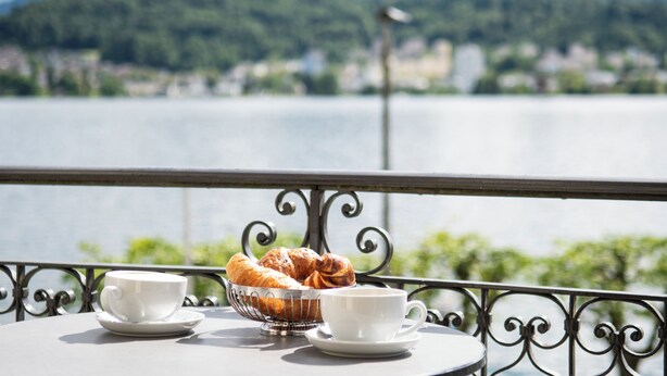 Une table avec un panier de croissants et deux tasses, donnant sur un lac avec des montagnes en arrière-plan.
