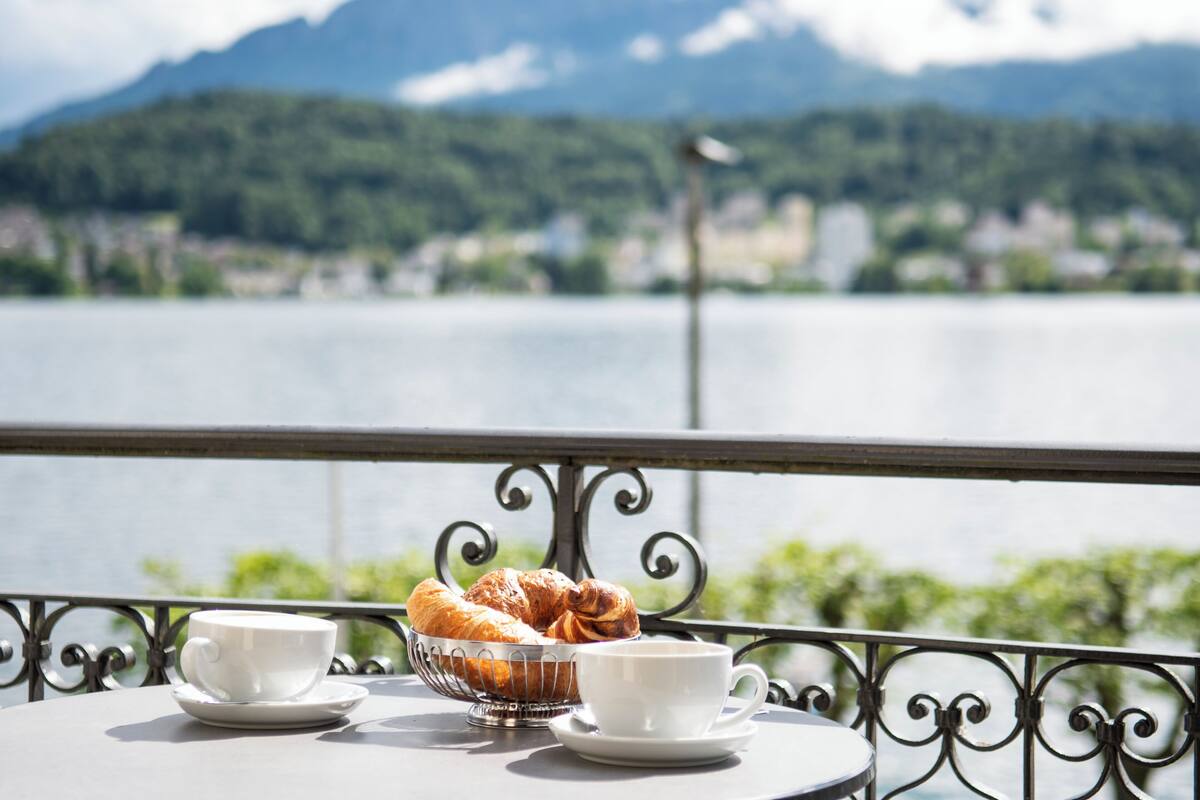 A table with a croissant basket and two cups, overlooking a lake with mountains in the background.