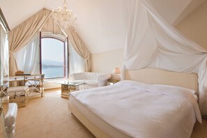 A bedroom with a large bed, a sitting area with a table and chairs, a view of the sea, and a chandelier. - Hotel Seeburg (Lucerne)