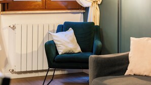 A room with a window, a radiator, a green armchair, a grey sofa, and a shelf with books and a bottle. - Pettinari Apartment (Roma)