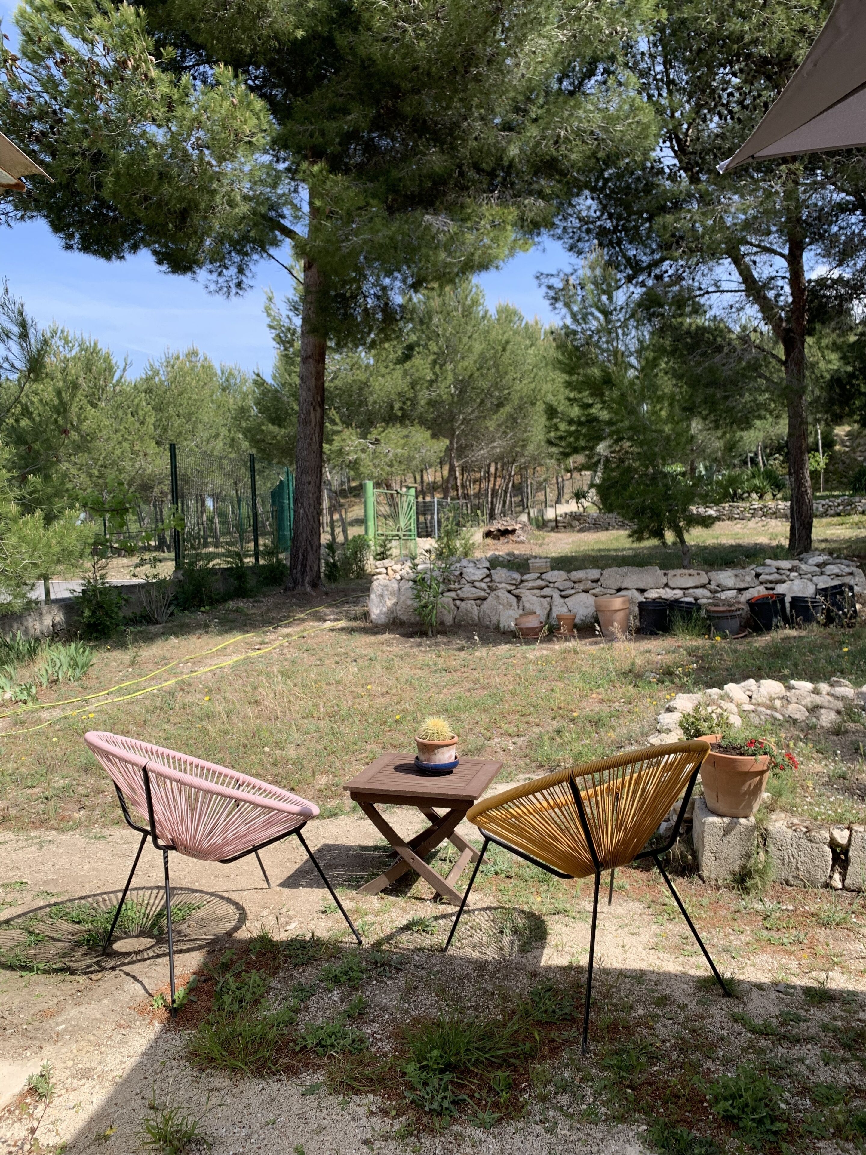 Une terrasse avec deux fauteuils en osier et une petite table, entourée d’arbres et d’un mur de pierre.