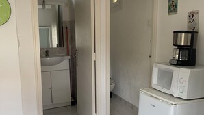 Een compacte keuken met een koelkast, magnetron en koffiezetapparaat. Een badkamer is zichtbaar door een geopende deur.