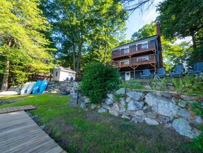 A cabin with a stone wall, a wooden deck, and a small garden area. - Leicester Lake Front Home, Come Relax! (Leicester)