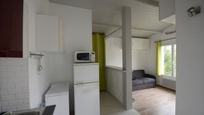 Una cocina compacta con microondas, un refrigerador pequeño y un lavamanos. En la zona de estar contigua, hay un sofá gris y una ventana con cortinas.