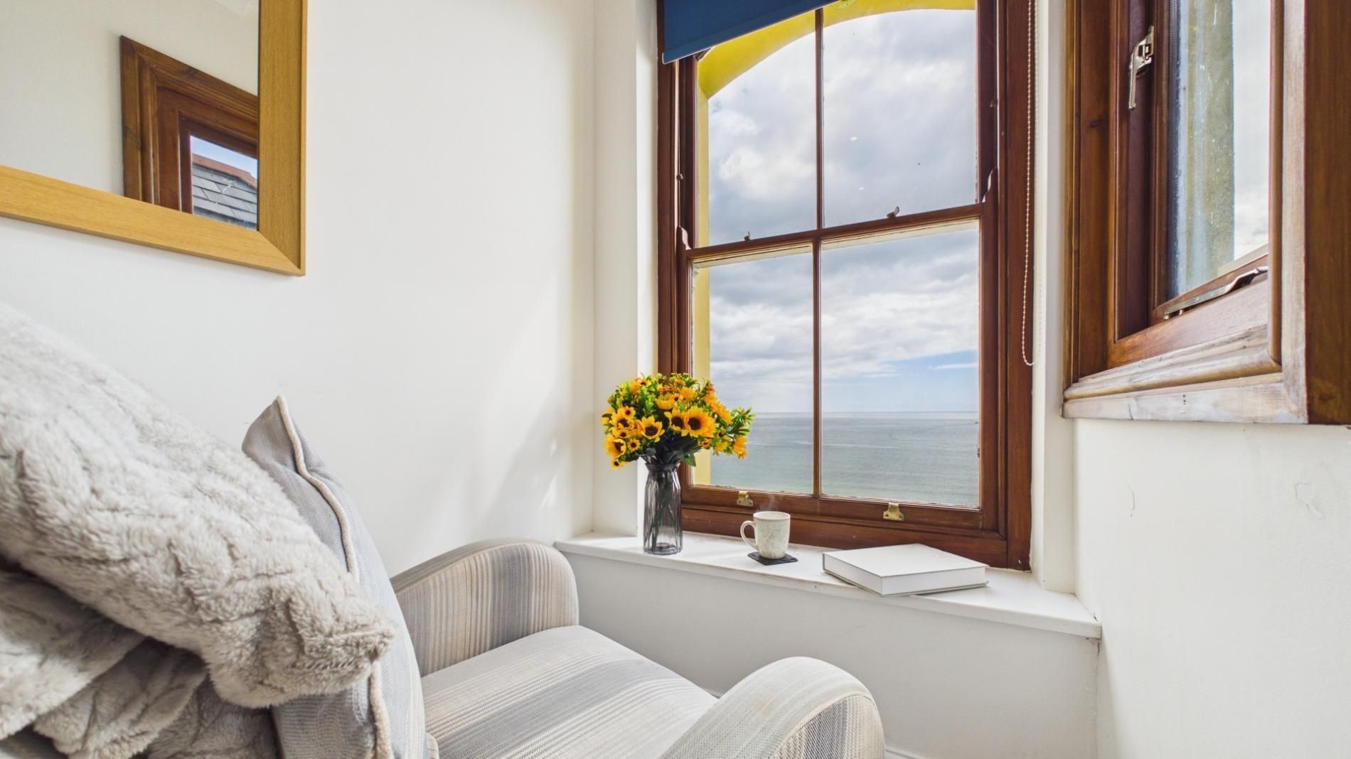 A room with a large window, a chair, a small table, and a vase with flowers.