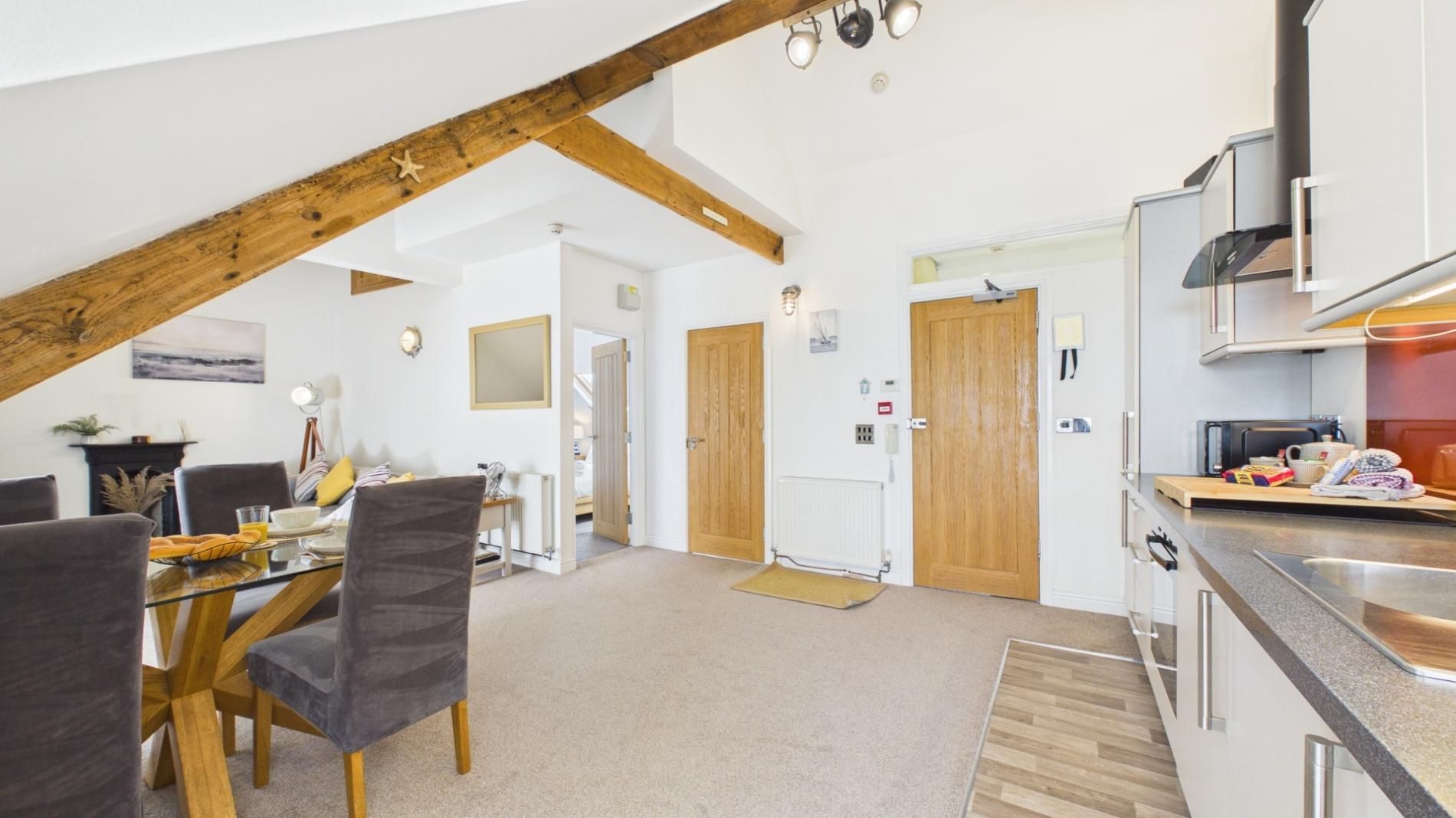 A modern kitchen with a wooden ceiling beam, a dining area with a table and chairs, and a view of the living room.