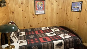 A wooden-paneled room with a bed covered in a patchwork quilt, a framed poster on the wall, and a table lamp. - Eagle Bay Cottage with Lake Rights (Eagle Bay)