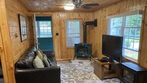 A cozy cabin interior with a leather sofa, a television, a fireplace, and a large window. - Eagle Bay Cottage with Lake Rights (Eagle Bay)