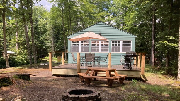 A rustic cabin with a deck, a picnic table, and a fire pit in a forest setting. - Eagle Bay Cottage with Lake Rights (Eagle Bay)