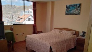 Une chambre à coucher comprenant un lit, une chaise, une petite table et une vue sur le paysage urbain, visible à travers une grande fenêtre.