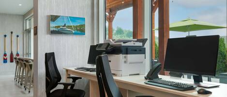 Un espacio de oficina moderno con un escritorio amplio, varias sillas, una impresora, un monitor de computadora y vistas al exterior a través de grandes ventanales.