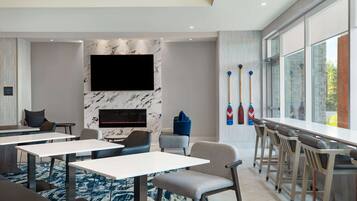 A modern hotel lobby with a flat-screen TV mounted on a marble wall, a bar area with stools, and a seating area with tables and chairs.