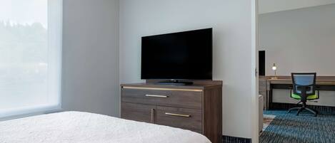 Quarto de hotel com uma cama, uma televisão em uma cômoda de madeira, uma escrivaninha com cadeira e vista para árvores pela janela.