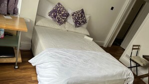 Ein ordentlich bezogenes Bett mit weißer Bettwäsche und zwei Zierkissen, ein Nachttisch aus Holz mit Fernbedienung und ein Sessel mit Sitzkissen.