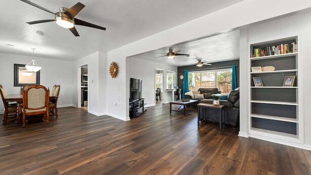 A spacious living room with hardwood floors, a dining area with a wooden table and chairs, and a bookshelf filled with books.