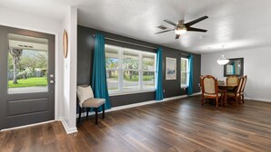 A spacious room with hardwood flooring, a dining area with a table and chairs, a ceiling fan, and large windows with curtains.