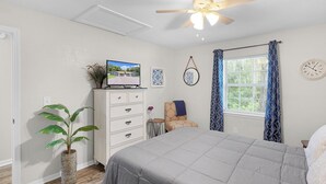 Ein Schlafzimmer mit einem Bett, einem Deckenventilator, einem Fernseher, einem Schminktisch, einer Pflanze, einem Sessel, einem Fenster mit Vorhängen und einer Wanduhr.