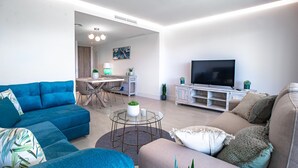 Un salon moderne avec un canapé bleu, un canapé gris et une table basse. On aperçoit un téléviseur posé sur un meuble blanc, et un coin repas à l’arrière-plan.