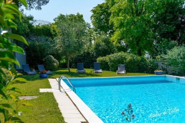 A swimming pool with a clear blue water, surrounded by green grass and trees. There are lounge chairs and a small garden area.