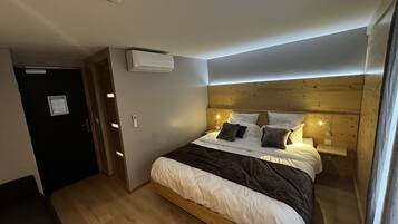 Un dormitorio con cabecera de madera, cama con ropa de cama blanca y oscura, unidad de aire acondicionado y mesita de noche con lámpara.