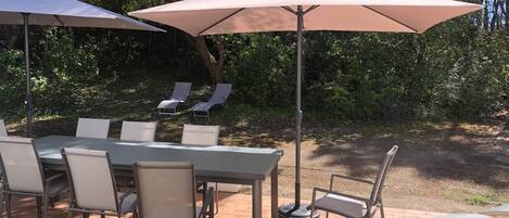 An outdoor patio with a table and chairs, two umbrellas, and a view of a wooded area.