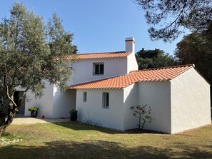A two-story building with a red-tiled roof, white walls, and a small garden area with a tree and potted plants. - Fully Equipped Villa—direct access to the beach, just a 3-minute walk away, and close to shops (L'Épine)