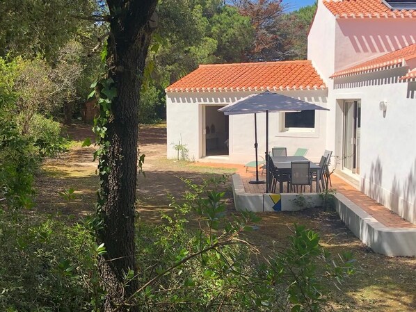 A white building with a red-tiled roof, a patio with a table and chairs, and a tree in the foreground. - Fully Equipped Villa—direct access to the beach, just a 3-minute walk away, and close to shops (L'Épine)