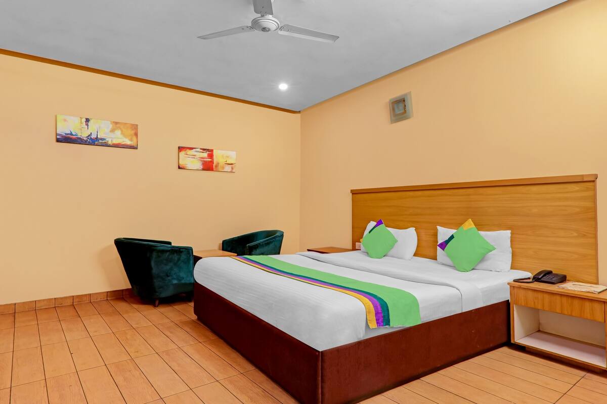Habitación de hotel con cabecera de madera, cama con ropa blanca y almohadas de colores, escritorio con teléfono y dos sillas.