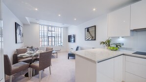 A modern kitchen with white cabinets, a marble countertop, and a dining area with a table and chairs. - Palace Wharf Apartments (London)