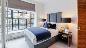 A modern bedroom with a large window, a bed with a blue blanket, two bedside tables with lamps, and a wooden door. - Palace Wharf Apartments (London)