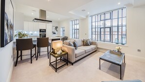 A modern living room with a sofa, chairs, a coffee table, and a kitchen in the background. - Palace Wharf Apartments (London)