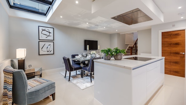 A modern kitchen with a central island, a dining area with a table and chairs, a built-in TV, and a staircase leading to an upper level. - Palace Wharf Apartments (London)