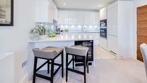 A modern kitchen with white cabinetry, a central island, two bar stools, and built-in appliances. - Palace Wharf Apartments (London)