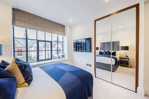 A bedroom with a large window, a bed with a blue and gold headboard, a bedside table with a lamp, and a wardrobe with a mirror.