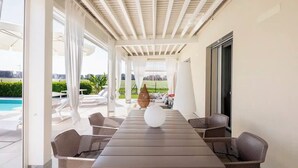 Un espace extérieur abrité, doté d’une longue table et de chaises, d’un vase décoratif, et offrant une vue sur une piscine et un espace verdoyant.