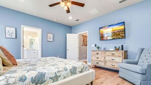 Un dormitorio con cama, cómoda, silla, ventilador de techo y televisor montado en la pared.