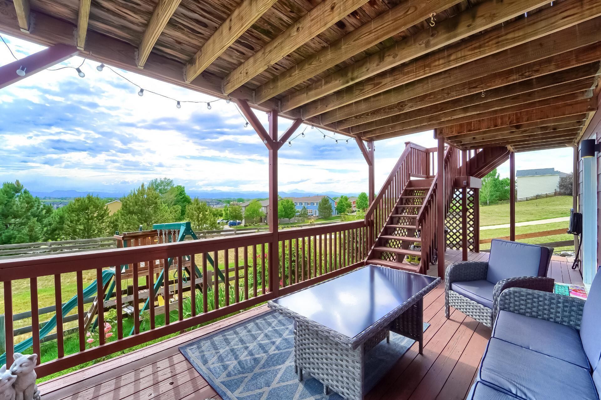A covered deck with a glass-top table and wicker furniture, overlooking a playground and a scenic landscape.